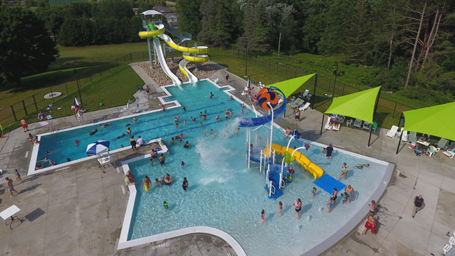 Overhead view of the outdoor Schulenburg Pool with zero depth beach entry, a water slide, and a giant play structure featuring dumping buckets